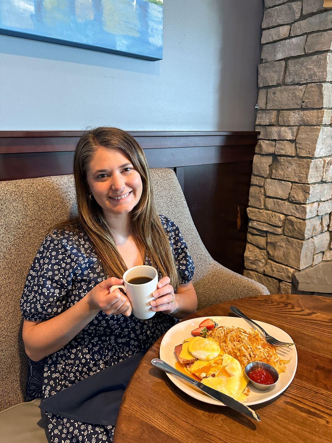 A person sits at a table posing with a cup of coffee and a plate of food at Plank