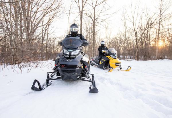 Two snowmobilers ride along a groomed trail as the sun hangs just above the horizon, casting a warm orange glow across the sky.