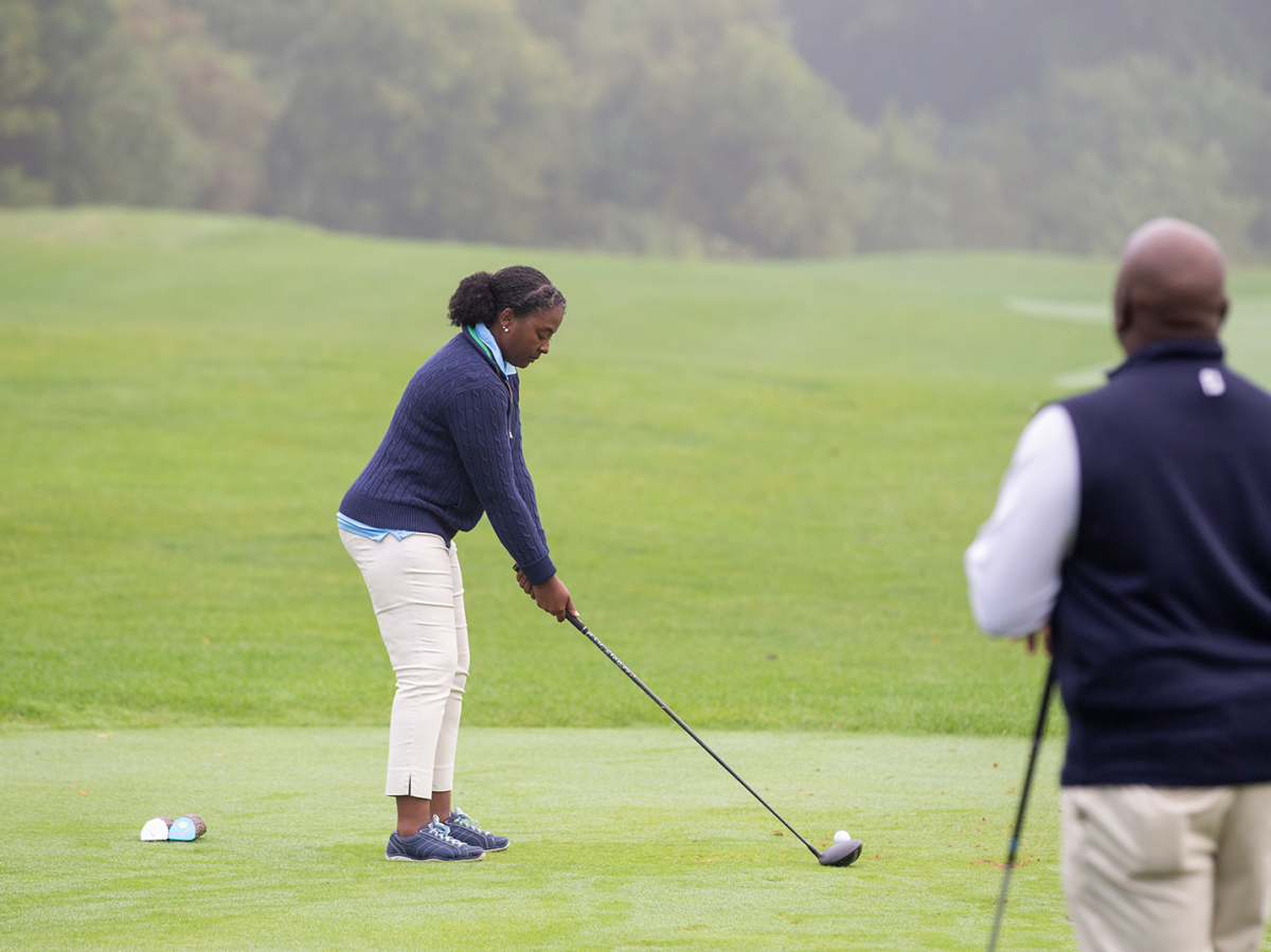 A woman golfer prepares to drive on a lush, green course, holding a club, while another person watches. The setting is calm with misty trees in the background.