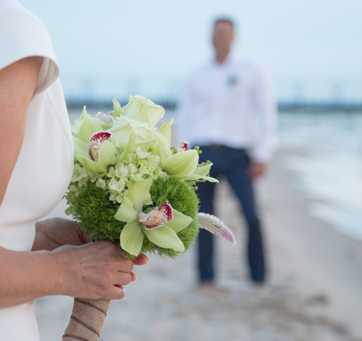 A bride walks along the beach holding flowers as she approaches the groom.