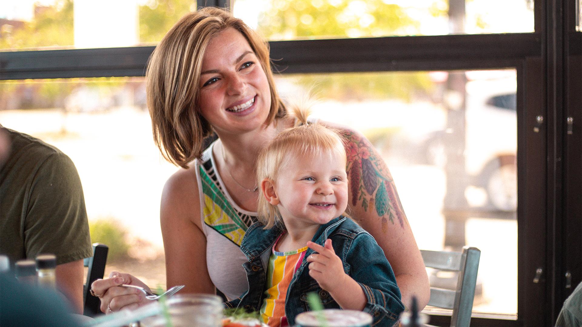 Smiling woman with short hair and colorful tattoo holds a happy toddler in a denim jacket at a bright, cheerful café table.