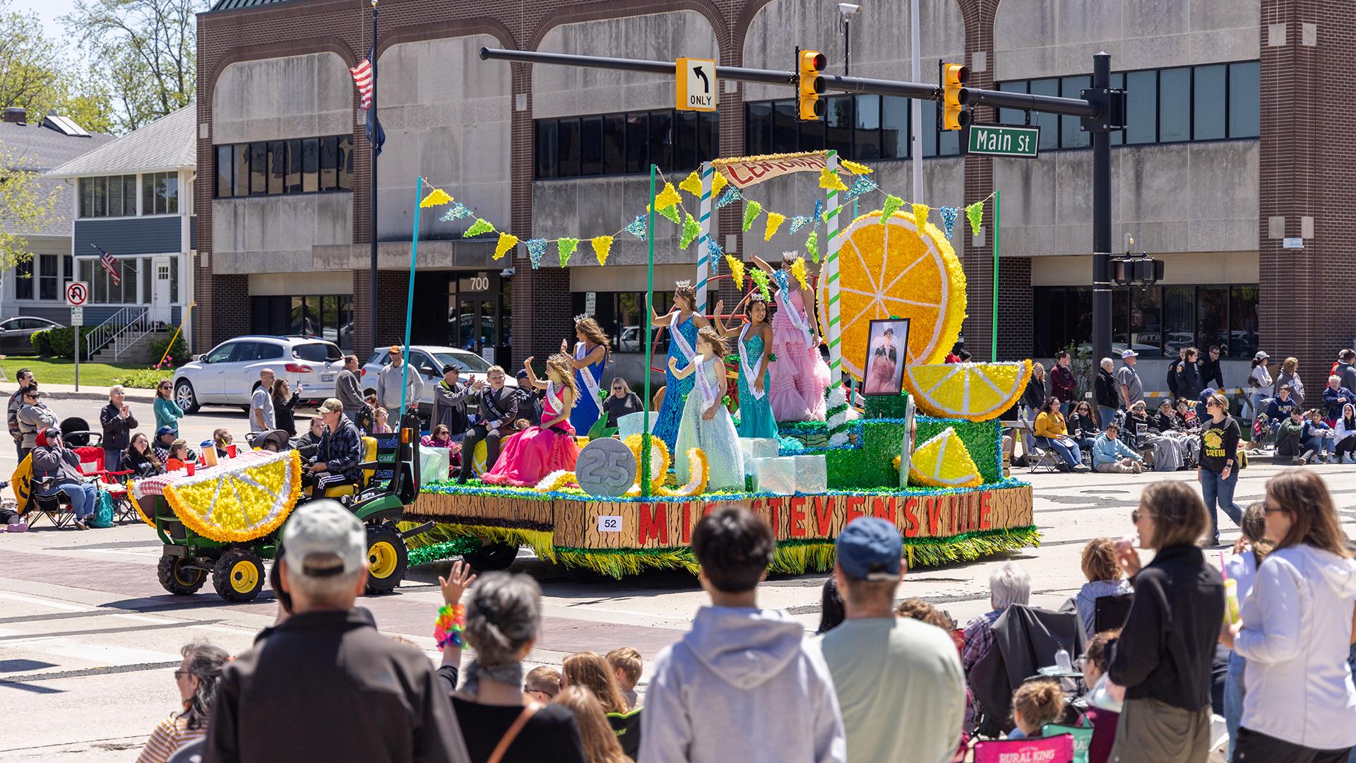 A vibrant Blossomtime parade float shaped like lemon slices carries people in colorful dresses waving. Spectators line the street, enjoying the sunny day.