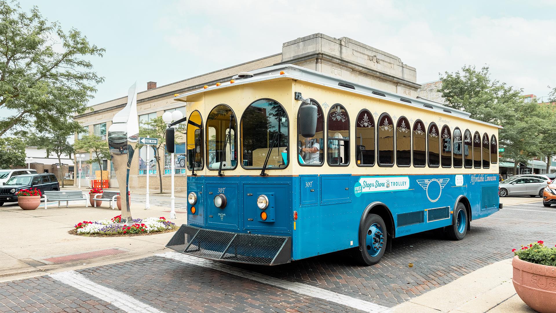 A trolley with arched windows drives through Downtown St. Joseph, surrounded by trees and flower beds create a lively charming feel.