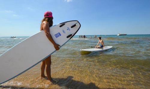 A close-up of a woman holding a paddleboard, preparing to join others on the water. In the background, a boy is already on a paddleboard and a group of people, some with boards, are gathered nearby.
