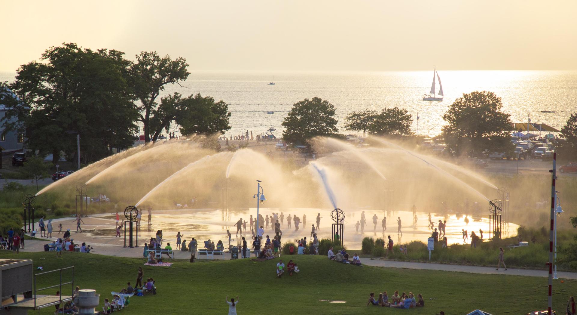 The Whirlpool Compass Fountain glows in the golden light of sunset with Lake Michigan and a sailboat visible in the distance.