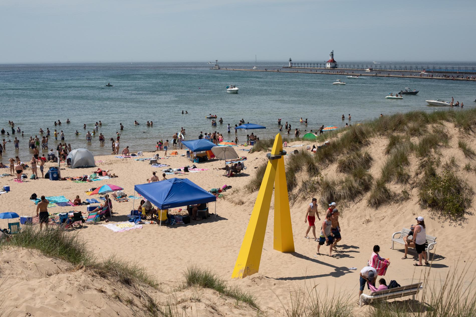 Silver Beach on a busy summer day. Yellow sculpture in the foreground, lots of people walking and seating in the sand, lighthouse in the background.