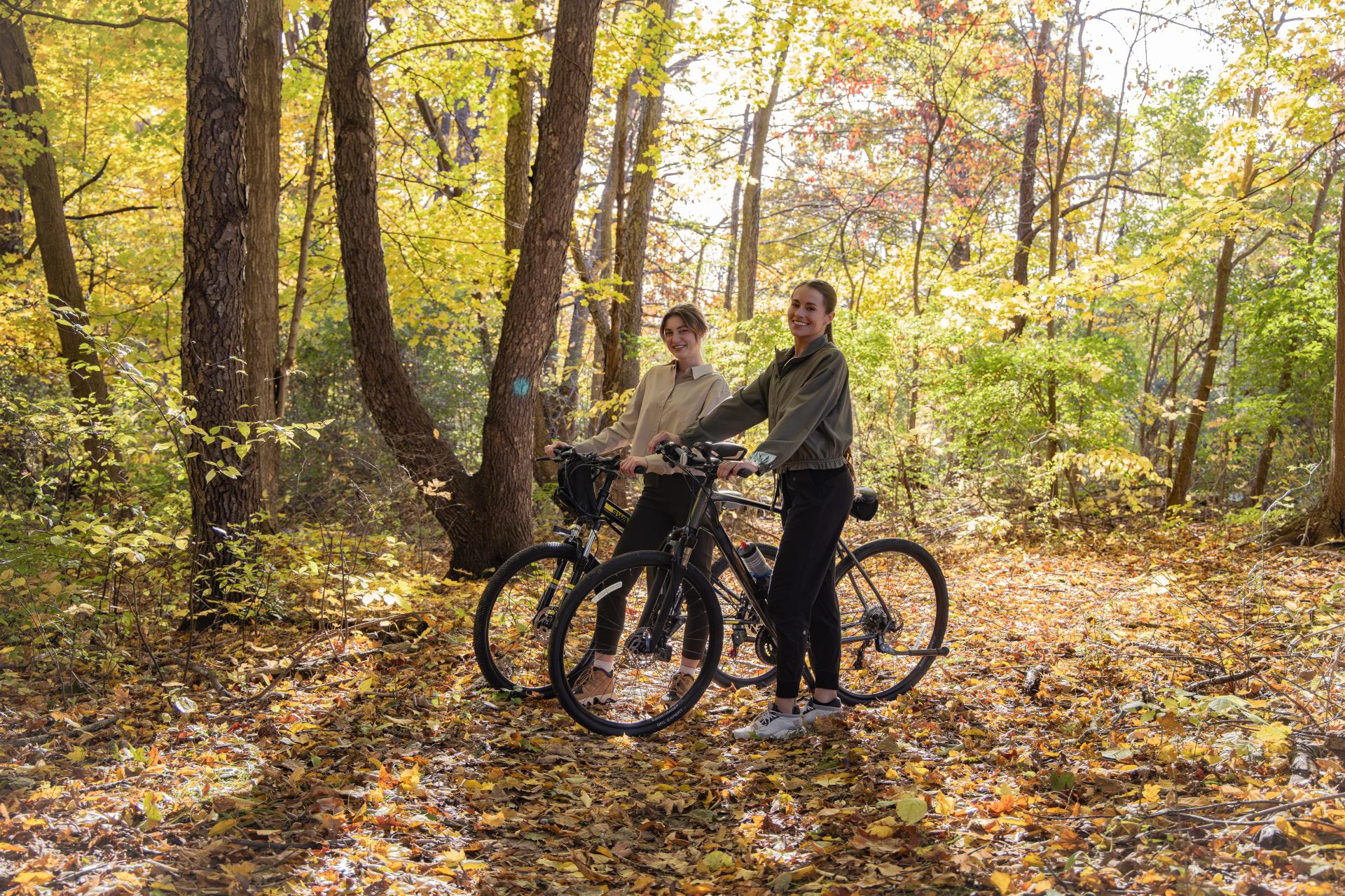 Two women stand with their mountain bikes beside a trail surrounded by vibrant fall colors.