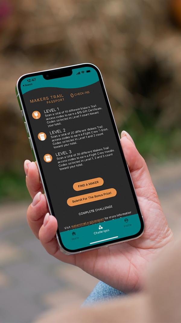 A hand holding up a phone displaying the Makers Trail Passport app